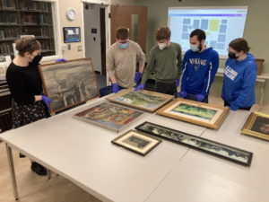 Madison stands holding a piece of artwork while four student assistants look on, examining additional pieces of artwork on a table 